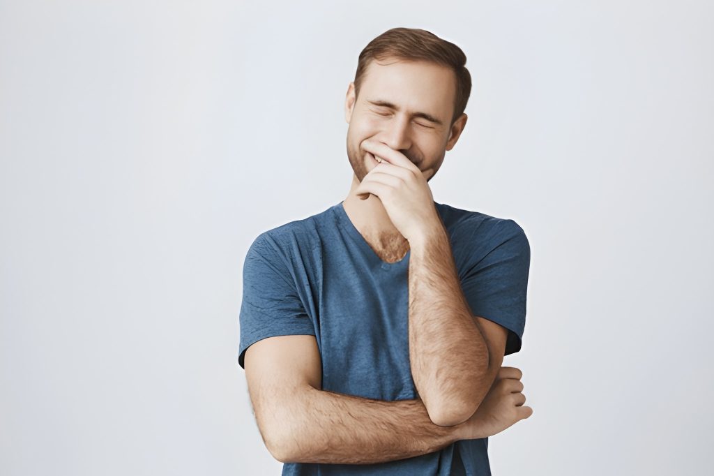 Homem a tampar parcialmente o sorriso com uma mão e olhos fechados